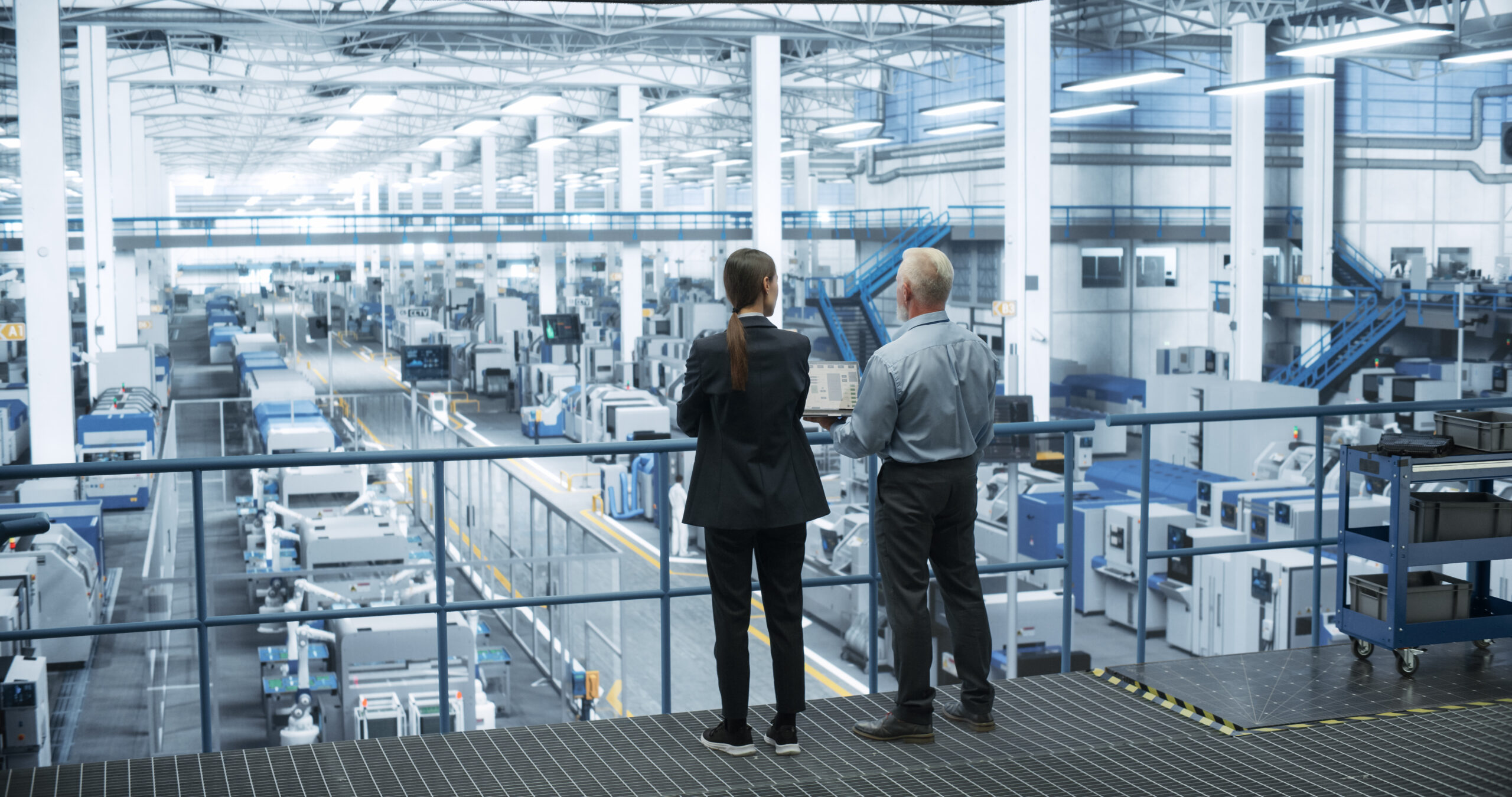Downloaded Middle Aged Engineer and Female Scientist Standing with Their Back, Using Laptop Computer and Talking in a Factory Facility with Equipment Producing Modern Technical Devices for Different Industries